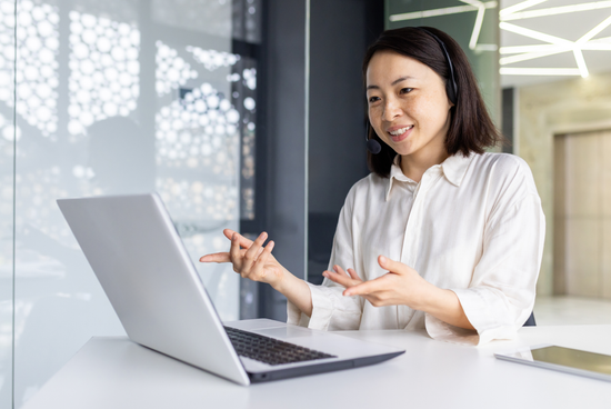 A woman in professional attire wearing a headset and smiling while gesturing during a video interview on her laptop in an office.