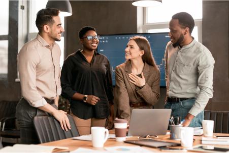 Group of smiling diverse professionals gathered around a laptop in an office setting, talking and collaborating with each other