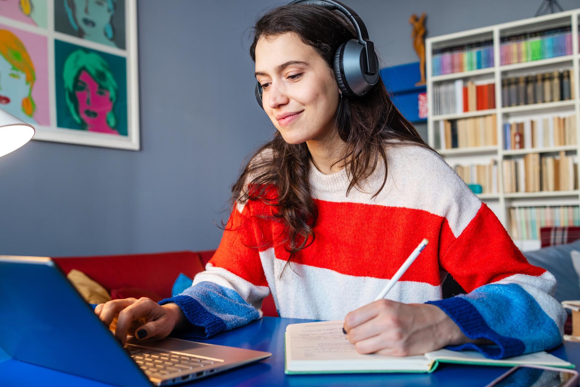 A person wearing headphones sits at a desk using a laptop and writing in a notebook, appearing focused while studying or applying for scholarships in a home setting with bookshelves in the background