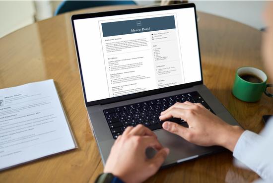 Person sitting at a desk using a laptop to create a resume with the help of an online resume writing services, with a cup of coffee and printed resume on the desk