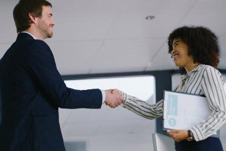 A woman holding a résumé smiles and shakes hands with a man in a business suit, connecting with a recruiter