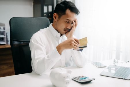 Stressed worker at a desk holding credit cards, looking down near a calculator, piggy bank, and laptop.