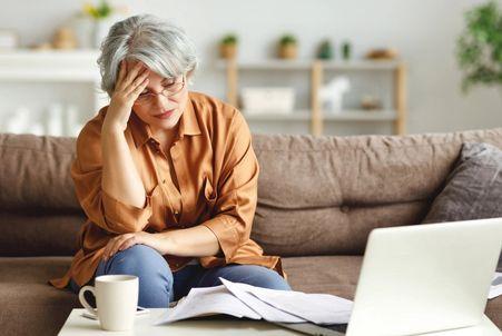 A mature professional holding her head in stress while analyzing household bills and retirement finances on a laptop.