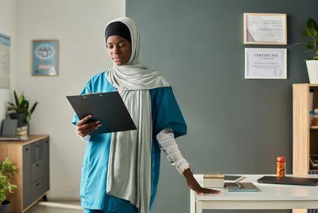 A healthcare provider in scrubs reviews a clipboard in her office, with professional certificates displayed on the wall.