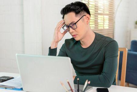 A job seeker holding their temple with a frustrated expression while sitting at a desk with a laptop, calculator, and papers.